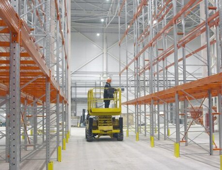 Mitarbeiter auf einer Hebebühne zwischen hohen Palettenregalen in einer Industriehalle.