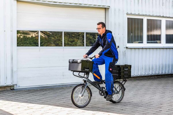 Mann in blau-schwarzer Arbeitskleidung fährt auf einem kleinen Transportfahrrad mit mehreren schwarzen Kisten vor einem hellen Industriegebäude.