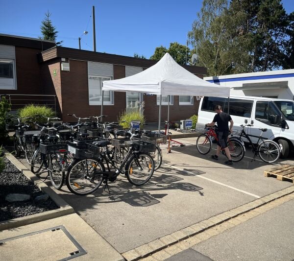 Mehrere Werksfahrräder stehen auf einem Parkplatz vor einem Gebäude. Neben den Werksfahrrädern befinden sich ein weißer Pavillon und ein weißer Transporter. Eine Person steht bei einem Fahrrad neben dem Fahrzeug.