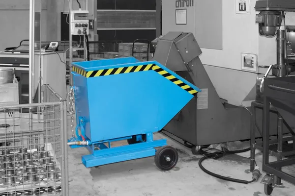Blauer Spänebehälter mit gelb-schwarzer Warnmarkierung und Fahrwerk in einer Produktionshalle, umgeben von Maschinen und Metallbehältern.