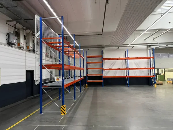 Leere Industriehalle mit blau-orangefarbenen Palettenregalen entlang der Wände. Gelb-schwarze Anfahrschutzwinkel sichern die Regalstützen am Boden.