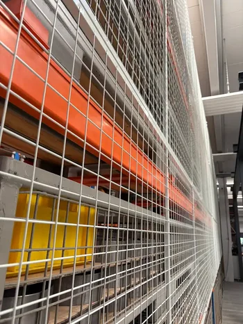 Nahaufnahme einer verzinkten Gitterrückwand an einem Palettenregal mit orangefarbenen Traversen und gelagerten Behältern in einer Industriehalle.