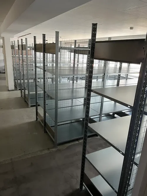 Mehrere leere Metallregale mit Gitterrückwänden und grauen Fachböden in einem Lagerraum.